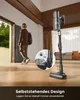 A person walks away from the camera into a doorway while a cordless vacuum cleaner stands upright in the foreground. Produktbild-Vorschau
