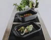 Two black roasting pans are displayed on a stovetop. One pan contains a cooked fish with herbs and vegetables, while the other contains roasted vegetables. Náhľad obrázku produktu