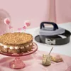 A cake is displayed on a pink cake stand, with a springform pan and a small jar of spread in the background, all set against a pink and white backdrop. Produktbild-Vorschau
