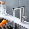 A modern kitchen faucet with water running into a sink, next to two glass bottles and a fruit basket. Produktbild-Vorschau