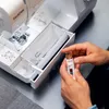 A person's hands are shown holding a sewing machine bobbin and bobbin case, with the sewing machine open in the background. Produktbild-Vorschau