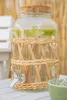 A glass beverage dispenser with a woven rattan-like sleeve and a silver spigot, filled with water and sliced green fruit, sits on a white table. A green bowl is partially visible in the foreground. Náhled obrázku produktu