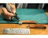 A person is using a Dremel rotary tool to engrave a name onto a leather keychain. Other leather keychains and a stencil are also visible. Anteprima dell’immagine prodotto