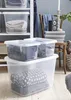 Storage boxes filled with folded clothes and blankets are stacked on a shelf and on the floor, with a chair and other household items in the background. Voir les images du produit