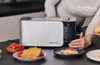 A person is using a toaster in a kitchen setting, with food items like waffles and tomatoes around. Voir les images du produit