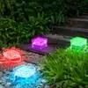 Four illuminated cube-shaped solar lights in red, purple, green, and blue are placed on a garden path among stones and plants. Produktbild-Vorschau