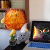 A plush sun toy sits on a desk next to a laptop displaying a solar system image, with a small potted plant nearby. Produktbild-Vorschau