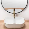 A white oval basin with a gold rim and a gold faucet is shown in a bathroom setting, with a round mirror reflecting the scene. Náhľad obrázku produktu