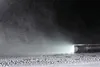 A black flashlight is lying on a surface covered in water droplets, with its beam illuminating the mist in front of it. Produktbild-Vorschau