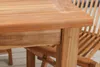 A close-up view of the corner of a wooden garden table and a chair in the background. Produktbild-Vorschau