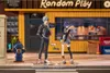 Two anime-style figures stand on a sidewalk in front of a building with a "Random Play" sign. Produktbild-Vorschau