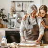 An older woman and a younger woman are in a kitchen, baking together. The older woman is rolling out dough with a rolling pin, while the younger woman has her arms around her. Produktbild-Vorschau