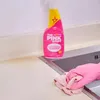 A pink spray bottle of cleaning product sits on a kitchen counter next to a sink, with a hand in a pink glove wiping the counter. Produktbild-Vorschau