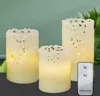 Three LED candles of varying heights are arranged on a dark surface, with a green background and foliage visible behind them. A remote control is placed in the foreground. Produktbild-Vorschau