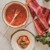 A top-down view of a meal preparation scene, featuring a cast iron pan filled with tomato sauce, a bowl of ravioli, a napkin, and fresh basil. Produktbild-Vorschau