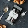 A gift box of salt and pepper spices is displayed on a dark, textured surface, surrounded by scattered peppercorns and a spoon of ground pepper. A spoon of salt and pieces of bread are also visible. Produktbild-Vorschau