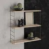 A three-tiered wall shelf with decorative items on a split concrete and black textured wall. Produktbild-Vorschau