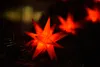 A close-up of a red star-shaped Christmas light decoration on a dark background, with other blurred lights in the distance. Produktbild-Vorschau