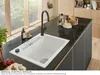 A modern kitchen sink area with a black granite countertop, a white sink, and a black faucet. There are also some kitchen items like soap, a brush, and food ingredients visible. Produktbild-Vorschau