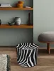 A zebra-print pouf sits on a wooden floor next to a rug and a gray ottoman, with shelves holding decorative items in the background. Produktbild-Vorschau
