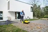 A man is using a yellow and black push sweeper to clear fallen leaves from a paved walkway in front of a modern white house. Produktbild-Vorschau