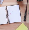 A close-up view of an open spiral notebook with a pocket containing a small card with colorful tabs. A dark brown wand-like object and a yellow notebook are also visible on a wooden surface. Produktbild-Vorschau