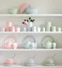 A white shelf displaying various ceramic tableware items in pastel colors. Produktbild-Vorschau