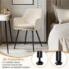 A beige velvet armchair with black metal legs is placed next to a white vanity table. A bed is visible in the background. Produktbild-Vorschau
