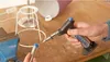 A person is using a Dremel VersaFlame butane torch to solder a metal component held in a vise, with parts of a lamp visible. Náhled obrázku produktu