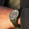 A close-up shot of a person's wrist wearing an olive green Ice-Watch with a black fabric strap. Voir les images du produit