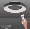 A hand holding a remote control points towards a modern LED ceiling lamp with a circular design. Produktbild-Vorschau