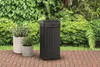 A black trash can sits on a brick patio next to two potted plants, with lush green foliage in the background. Anteprima dell’immagine prodotto