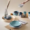 A collection of tableware, including bowls, mugs, and plates, arranged on a table with a decorative grass stalk in a vase. Produktbild-Vorschau