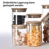 Several glass storage jars with wooden lids are stacked and arranged, filled with different food items. Produktbild-Vorschau