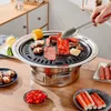 A hand is using tongs to place a piece of meat on a round Korean BBQ grill, which is set up on a wooden table. Various food items like sausages, mushrooms, corn, and crab sticks are also visible around the grill. Náhled obrázku produktu