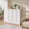A white cabinet with drawers and doors is shown in a room setting, next to a chair and plants. Produktbild-Vorschau
