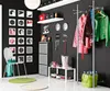 A modern entryway or hallway with a black wall, featuring a white chair with a green scarf, a coat rack with several jackets, a shoe rack with boots and shoes, and decorative elements on the wall. Produktbild-Vorschau