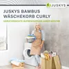 A woman is standing next to a washing machine and a laundry basket, holding a basket of laundry. Produktbild-Vorschau