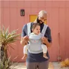 A man is holding a baby in a carrier in front of a pink building. Produktbild-Vorschau