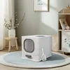 A white cat litter box with a gray entrance and scoop is placed on a round blue rug in a room with a shelf and a small stool with a plant. Podgląd zdjęcia produktu
