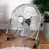 A chrome floor fan is placed on a wooden floor next to a potted plant and a chair. Voir les images du produit