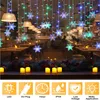 A festive display of snowflake string lights with colorful LEDs, set against a blurred background of a restaurant interior. Several LED candles are placed on a windowsill in the foreground. Produktbild-Vorschau