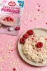 A bowl of porridge with raspberries and white chocolate drizzle, next to a product package, on a pink background with scattered oats and raspberry pieces. Produktbild-Vorschau