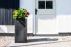 A tall, rectangular planter with a flowering plant sits on a paved walkway in front of a white brick house with a white door. Produktbild-Vorschau