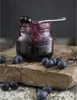 A jar of blueberry jam with blueberries scattered around it on a wooden surface. Produktbild-Vorschau