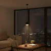 A dimly lit living room at night with a large window overlooking a city skyline. A pendant light hangs over a coffee table with a book, glasses, and a cup of coffee. Produktbild-Vorschau