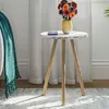 A small round side table with a marble-like top and wooden legs, holding a yellow vase with colorful flowers and a small blue container. A teal blanket is draped over a nearby chair. Produktbild-Vorschau
