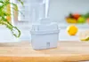 A water filter cartridge sits on a wooden cutting board, with a blurred background of a pitcher and some fruit. Produktbild-Vorschau