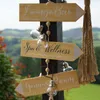 A wooden signpost with three directional signs pointing to 'Lounge Bar', 'Spa & Wellness', and 'Genuss Raum', decorated with string lights and wooden beads, set against a blurred outdoor landscape. Produktbild-Vorschau