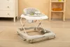 A baby walker is placed on a rug in a room with a dresser and shelves in the background. Podgląd zdjęcia produktu